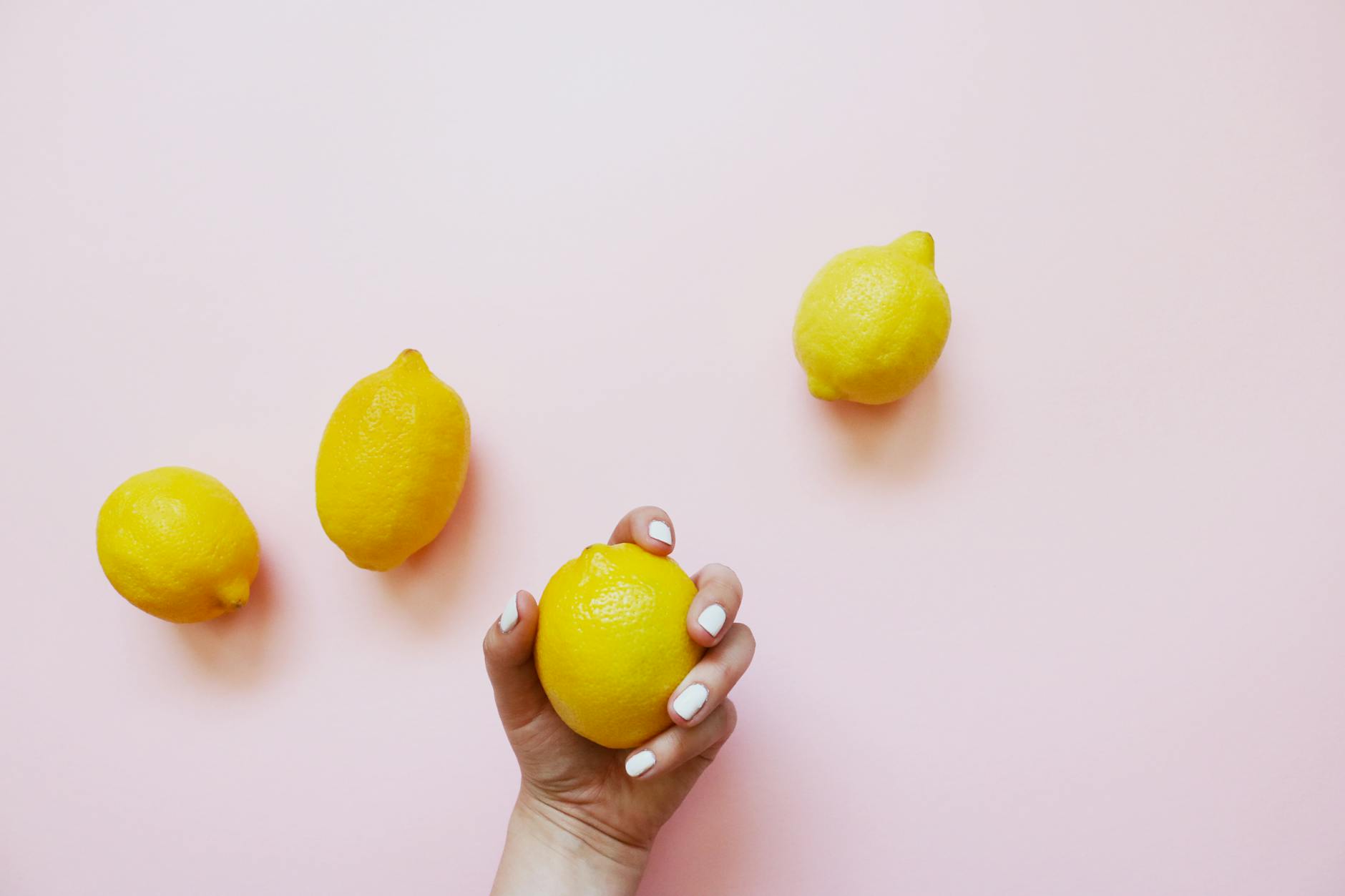 A hand holding a fresh lemon on a soft pink background surrounded by more lemons, symbolizing the design of lemon clitoral vibrators.