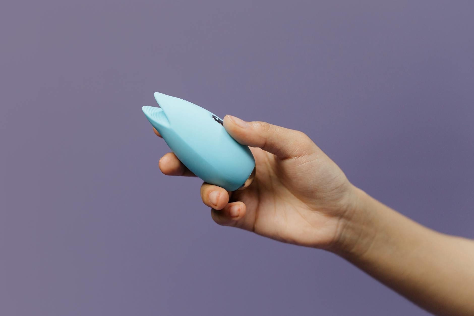 Hand holding a blue silicone clitoral vibrator against a purple background